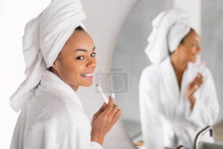 Téléchargez les photos : Soins dentaires. Heureuse femme noire tenant une brosse à dents prend soin de ses dents dans un cadre de salle de bain moderne, se préparer et prendre soin de l'hygiène buccodentaire le matin, mettant l'accent sur la beauté sourire naturel - en image libre de droit