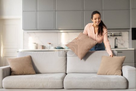 Photo for Smiling woman in pastel shirt arranging soft pillows on modern grey couch, adding touch of coziness to the bright, well-designed living room, free space - Royalty Free Image