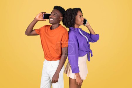 Photo for Side by side, a young black man in an orange shirt and a young woman in a purple blouse happily talk on their smartphones, back-to-back, against a vivid yellow backdrop, studio - Royalty Free Image