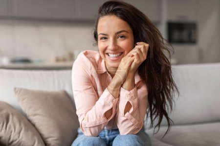 Photo for Radiant woman with lustrous hair and peach blouse exudes happiness, resting her chin on her hands while seated on comfortable couch at home - Royalty Free Image