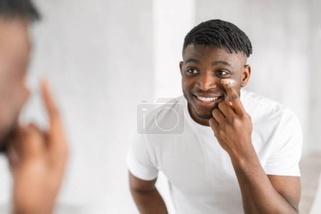 Photo for Cheerful millennial black guy applying moisturizer on face smiling to his reflection in mirror, enjoys his morning routine of pampering and selfcare in modern bathroom interior. Free space - Royalty Free Image
