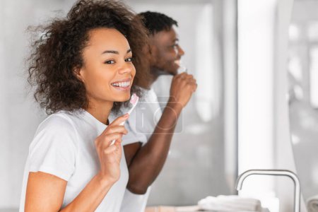 Téléchargez les photos : Portrait de jeunes couples afro-américains brossant à dents pendant leur routine matinale, souriant alors qu'ils s'engagent dans un moment de soins dentaires, debout ensemble dans une salle de bain moderne. Concentration sélective - en image libre de droit