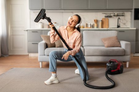 Photo for Playful woman cleans at home while listening to music on headphones, creatively mimicking playing guitar with her vacuum cleaner in living room interior - Royalty Free Image