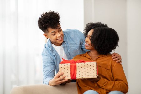 Téléchargez les photos : Délicieuse femme afro-américaine millénaire avec un large sourire reçoit une boîte-cadeau à pois avec un arc rouge d'un homme, profitant tous deux d'un moment agréable et chaleureux ensemble dans une pièce lumineuse - en image libre de droit