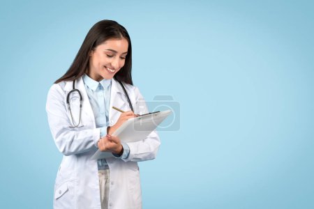 Engaged young female doctor writing on clipboard with smile, wearing lab coat and stethoscope, standing against light blue background, free space