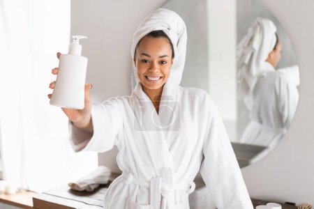 Téléchargez les photos : Heureuse jeune femme noire montrant bouteille distributeur recommandant produit cosmétique debout dans le peignoir avec serviette enveloppée sur la tête dans la salle de bain, souriant à la caméra. S'occuper de soi et prendre soin de soi - en image libre de droit