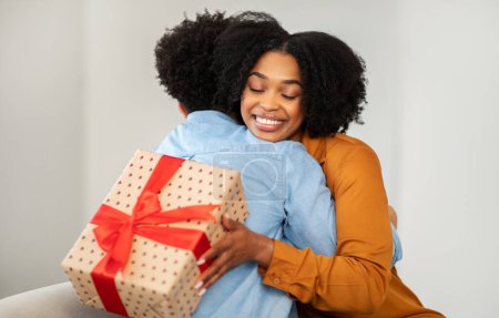 Photo for Radiant African American millennial woman with a bright smile hugs a man while holding a gift wrapped in polka-dotted paper with a red bow, expressing gratitude and affection - Royalty Free Image