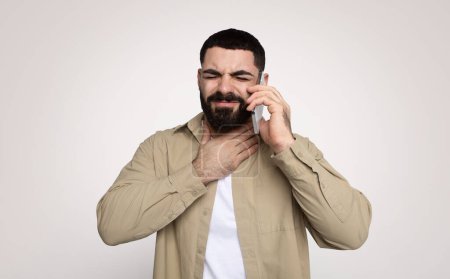 Photo for Man with a beard appears distressed while talking on the phone, hand on throat suggesting difficulty speaking or a sore throat, in a beige shirt against a plain background - Royalty Free Image
