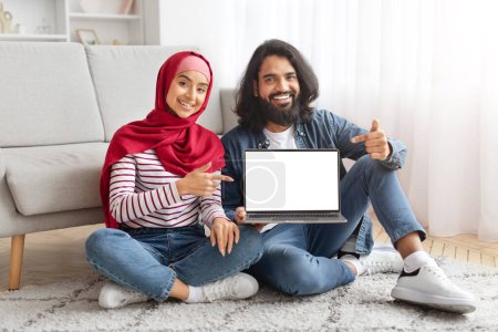 Photo for Young muslim couple pointing fingers at laptop with blank white screen, sitting together on floor in living room at home, smiling arab man and woman in hijab recommending new website, mockup - Royalty Free Image