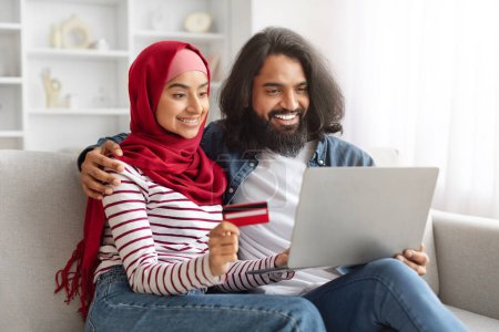 Téléchargez les photos : Paiements faciles. Heureux jeune couple musulman avec ordinateur portable et carte de crédit se détendre sur le canapé à la maison, joyeux époux islamiques faisant des achats en ligne, profiter des achats sur Internet, Copier l'espace - en image libre de droit