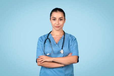 Competent young female nurse with arms crossed, wearing blue scrubs and stethoscope, exuding professionalism and readiness on blue background