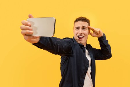 Téléchargez les photos : Charismatique beau jeune homme blond prenant selfie sur smartphone, gestuelle et grimaçante à la caméra téléphonique, isolé sur fond jaune. Influenceur fabrication de contenu, streaming - en image libre de droit