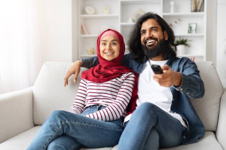 Foto de Retrato de feliz pareja musulmana viendo televisión y divirtiéndose en casa juntos, jóvenes cónyuges árabes relajándose en el sofá en la sala de estar, hombre sosteniendo el mando a distancia y señalando en la pantalla - Imagen libre de derechos