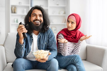 Angry muslim woman looking at husband watching tv at home, unhappy arabic lady in hijab female feeling lonely, excited smiling man holding remote controller and ignoring his upset girlfriend, closeup