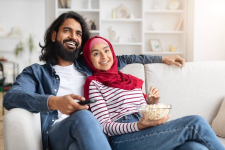 Foto de Relajada pareja musulmana descansando en el sofá con un tazón de palomitas de maíz, feliz hombre y mujer árabe sonriente en hijab compartiendo un momento acogedor juntos, viendo la televisión y descansando en el acogedor interior de la sala de estar - Imagen libre de derechos