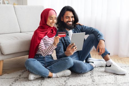 Téléchargez les photos : Concept d'achat en ligne. Couple musulman heureux utilisant la tablette numérique et la carte de crédit à la maison, conjoints arabes avec Gadget moderne achetant des choses dans Internet tout en étant assis sur le sol dans le salon - en image libre de droit