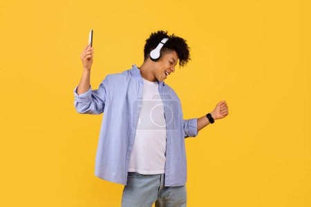 Photo for Joyful young black guy with curly hair dances to the rhythm in his headphones, smartphone in hand, showcasing the universal language of music on vibrant yellow background - Royalty Free Image
