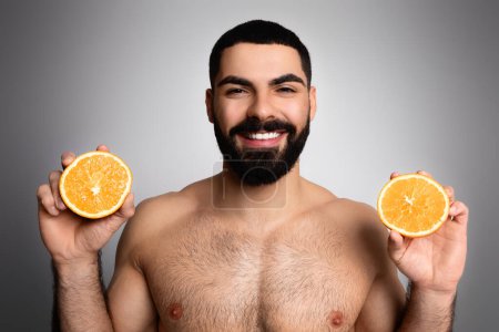Téléchargez les photos : Vitamine C dans la routine de soins de la peau pour les hommes. Beau beau mec arabe à moitié nu montrant des moitiés orange à la caméra et souriant, posant isolé sur fond de studio gris, gros plan - en image libre de droit