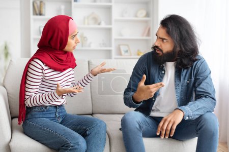 Foto de Problemas de relación. Retrato de una joven pareja musulmana discutiendo en casa, cónyuges árabes milenarios sentados en el sofá y culpándose mutuamente, sufriendo malentendidos y crisis matrimonial - Imagen libre de derechos