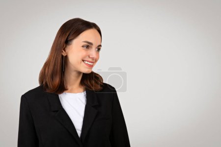 Foto de La confiada empresaria caucásica milenaria con una cálida sonrisa vistiendo un traje negro clásico mira a un lado, retratando la accesibilidad y el profesionalismo sobre un fondo silencioso - Imagen libre de derechos