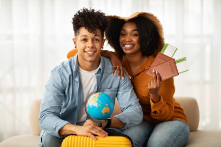 Foto de Feliz pareja afroamericana milenaria con pasaportes y tarjetas de embarque listas para viajar, sentadas al lado de un globo y una maleta de color amarillo brillante, irradiando emoción por una nueva aventura - Imagen libre de derechos