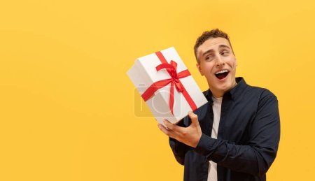 Photo for Excited young guy holding white gif box with red bow next to his ear, wondering what is inside, isolated on yellow background. Birthday man enjoying his presents, panorama with copy space - Royalty Free Image