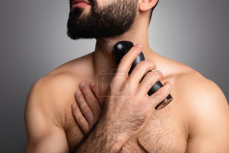 Téléchargez les photos : Coupé de jeune homme musclé coupant sa barbe épaisse ou son cou rasé avec un rasoir électrique, isolé sur fond de studio gris. Concept d'outils de soin de la barbe - en image libre de droit