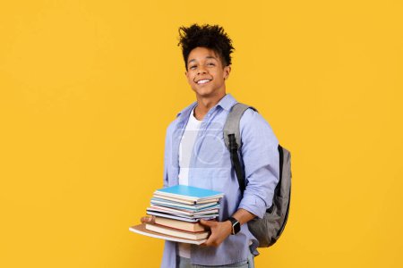 Foto de Alegre estudiante negro llevando mochila y pila de libros, sugiriendo preparación para desafíos académicos, en un animado fondo amarillo - Imagen libre de derechos