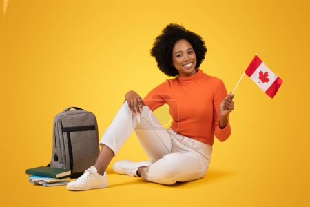 Photo for A friendly young woman with a radiant smile and curly hair sits comfortably on the floor, holding a Canadian flag, with school supplies and a backpack nearby on a yellow background - Royalty Free Image
