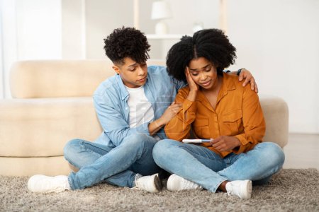 Compassionate African American man comforting a concerned woman, both seated on the floor, with the mans arm around her as she holds a tablet, in a serene home environment