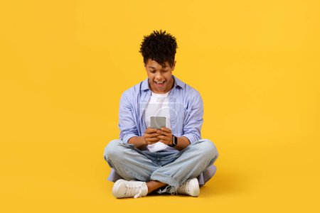 Entzückter Schwarzer, der in sein Smartphone vertieft ist, im Internet surft oder in den sozialen Medien scrollt, im Schneidersitz mit strahlendem Lächeln vor leuchtend gelbem Hintergrund