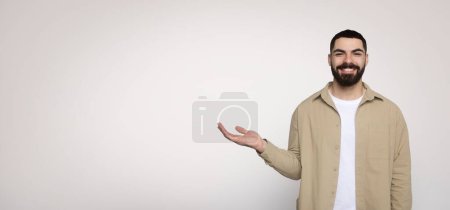 Téléchargez les photos : Affable homme barbu souriant et gesticulant la main ouverte comme tenant quelque chose d'invisible, vêtu d'une chemise beige décontractée et d'un t-shirt blanc, sur un large fond clair - en image libre de droit