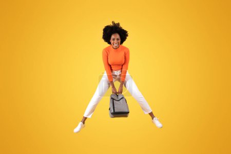 Téléchargez les photos : Plan dynamique d'une jeune femme afro-américaine ravie aux cheveux bouclés, tenant un sac à dos entre les jambes, vêtue d'une chemise orange et d'un pantalon blanc sur fond jaune - en image libre de droit