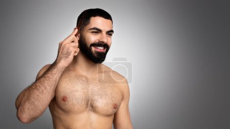 Hair care, healthy hair growth for men, beauty treatment concept. Happy handsome half-naked arab man combing his hair with brush and smiling, looking at copy space, grey background
