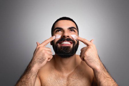 Cheerful handsome bearded millennial middle eastern man using face cream and smiling, moisturizing skin after shower, posing topless over grey studio background, looking up at copy space above him
