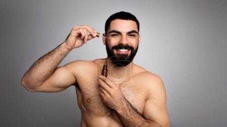 Photo for Good-looking cheerful bearded athletic half-naked young middle eastern applying anti-aging serum or oil on his cheek, looking at camera, skin care advert, grey background, panorama with copy space - Royalty Free Image