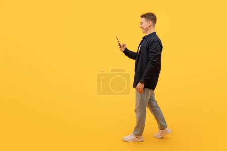 Foto de Vista lateral del chico rubio en casual con teléfono inteligente en la mano caminando hacia el espacio en blanco de copia, mensajes de texto amigos, aislado en el fondo del estudio amarillo, longitud completa. Comunicación digital - Imagen libre de derechos