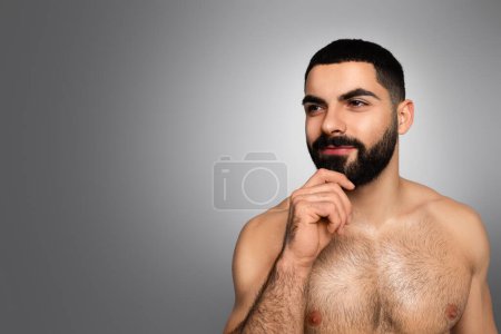 Foto de Concepto de acicalamiento de barba. Atractivo chico árabe milenario tocando su bonita barba y mirando el espacio en blanco de copia para la publicidad, posando en topless sobre fondo gris estudio - Imagen libre de derechos