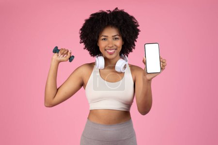 Téléchargez les photos : Enthousiaste jeune femme noire multitâche en levant haltère et en affichant smartphone avec écran blanc, combinant fitness et technologie, fond rose, maquette - en image libre de droit