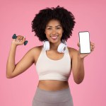 Enthusiastic young black woman multitasking by lifting dumbbell and displaying smartphone with blank screen, combining fitness and technology, pink background, mockup