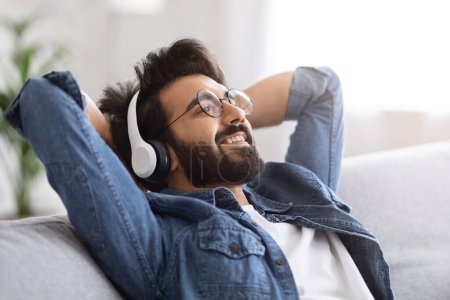 Photo for Joyful bearded indian man wearing glasses and wireless headphones reclining on sofa at home, happy young eastern male enjoying music in relaxed, cozy living room atmosphere, closeup - Royalty Free Image