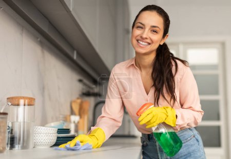 Téléchargez les photos : Femme amicale avec un sourire rayonnant, portant des gants jaunes, nettoyant son comptoir de cuisine avec un chiffon bleu et une bouteille de pulvérisation verte, appréciant la routine domestique - en image libre de droit