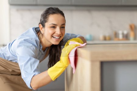 Téléchargez les photos : Focused and smiling woman wearing yellow gloves cleaning kitchen furniture with pink cloth, showing satisfaction in her domestic work - en image libre de droit