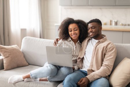 Photo for Online movie time. Happy black young spouses sitting with laptop at home, married couple spending quality time together, smiling and watching film on computer seated on sofa indoor - Royalty Free Image