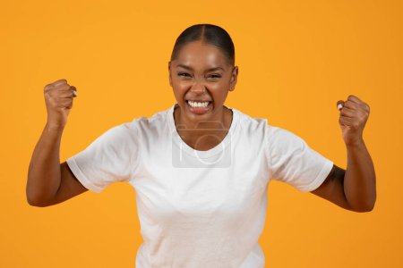Téléchargez les photos : Jeune afro-américaine dans les regards occasionnels dans la caméra avec une expression passionnée, serre ses poings dans la colère et l'effort, mettant en valeur la force et la détermination sur fond jaune, plan studio - en image libre de droit