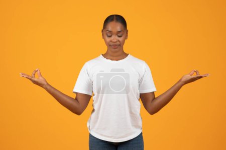 African american millennial lady meditating making om gesture, practices yoga in studio against orange background, standing with eyes closed in relaxed meditation pose. Mindfulness and relaxation