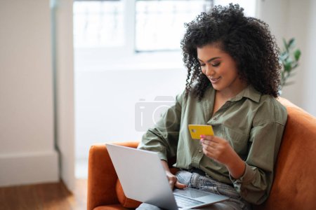 Téléchargez les photos : Jeune femme moderne avec un ordinateur portable et carte de crédit à la main se livre à des achats sur Internet dans le confort de sa maison. Acheteur navigue sur le site faisant l'achat et le paiement en ligne - en image libre de droit