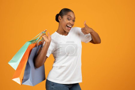 Foto de Joven mujer afroamericana sosteniendo bolsas de comprador y dando pulgares hacia arriba, expresando alegría con una sonrisa en el entorno del estudio amarillo. Compras aprobando los precios con descuento del Viernes Negro - Imagen libre de derechos