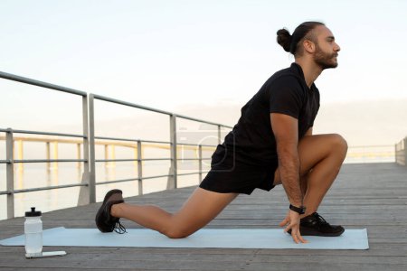 Side view of healthy sporty man flexing legs in a kneeling hip flexor stretch exercising outdoors. Athlethic guy enjoying his summer fitness routine by the sea pier, working on muscles flexibility