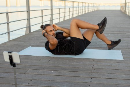 Téléchargez les photos : Homme sportif européen en vêtements de sport fait des exercices abdominaux sur la jetée et en utilisant le téléphone pour le streaming et le tournage de son entraînement en plein air au bord de la mer. Fitness blog et concept de bien-être - en image libre de droit